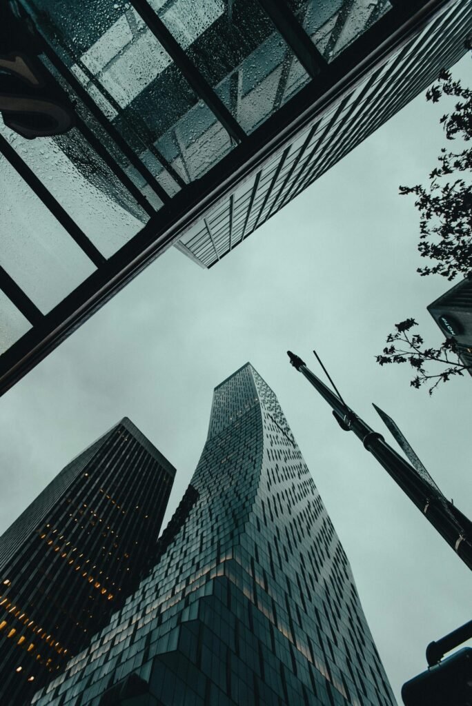 About Low-angle view of modern glass skyscrapers rising toward an overcast sky, framed by surrounding buildings and street elements.