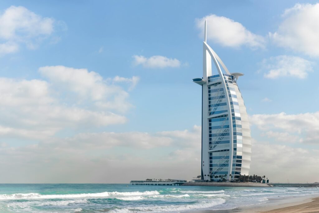 Burj Al Arab luxury hotel in Dubai standing on an artificial island with waves along the beach under a clear blue sky.