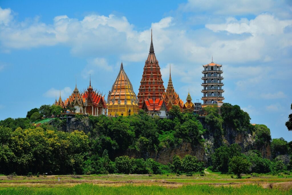 Destination Colorful Buddhist temple buildings and pagodas perched on a green hill, rising above fields and trees under a bright blue sky.