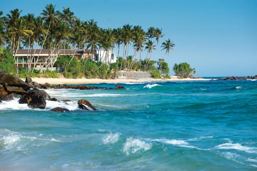 Tropical beach with turquoise ocean waves, palm trees, and seaside houses under a clear blue sky.