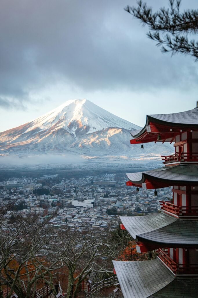 Mount Fuji overlooking a Japanese city with a traditional red pagoda in the foreground under a cloudy sky.