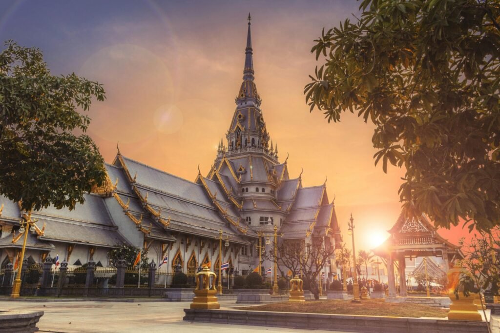 Traditional Thai temple complex at sunset with ornate rooftops, golden details, and a tall central spire framed by trees.